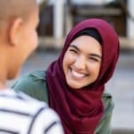 Cheerful woman in hijab smiling while talking to friend.